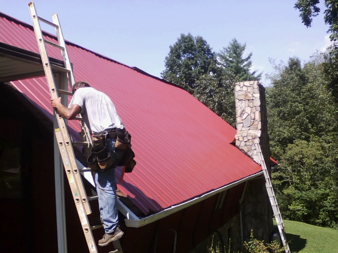 Roofer on a ladder beside a red metal roof and stone chimney — Soffit & Fascia Repair by Roofing Brothers in Fairdale